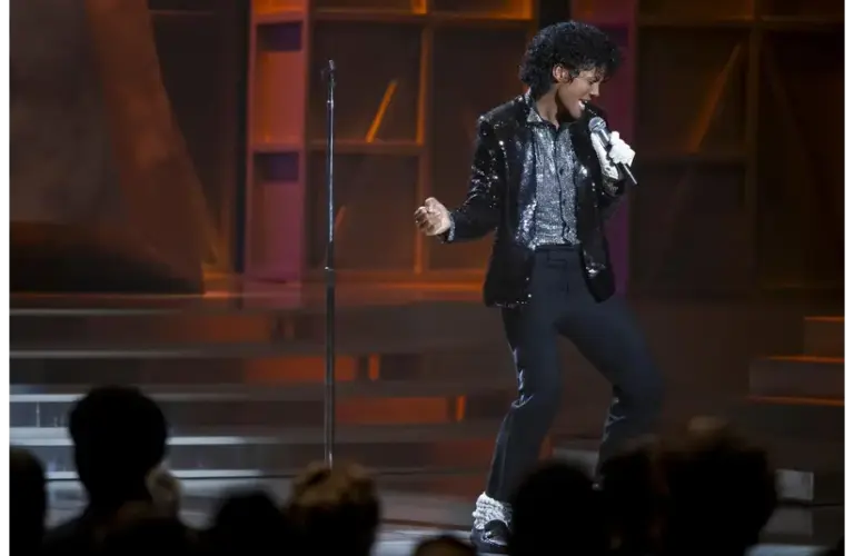 Jaafar Jackson in "Michael (2026)" performs on stage in a sparkling black jacket and glove, singing passionately into a microphone. The audience watches intently, creating an energetic atmosphere.