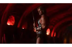 Nicholas Galitzine in Masters of the Universe (2026), dressed in a bronze-toned, gladiator-style outfit, wields a sword in a dimly lit cavern. Red light casts a dramatic, intense ambiance.