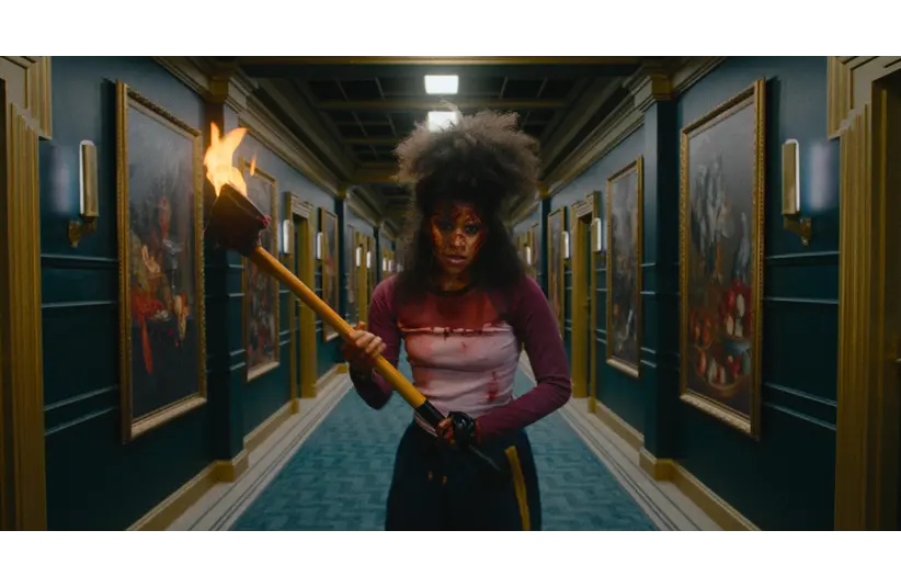 Zazie Beetz  with wild hair and face paint stands in a dim hallway holding a burning torch. Framed paintings line the walls, creating a tense, dramatic ambiance.