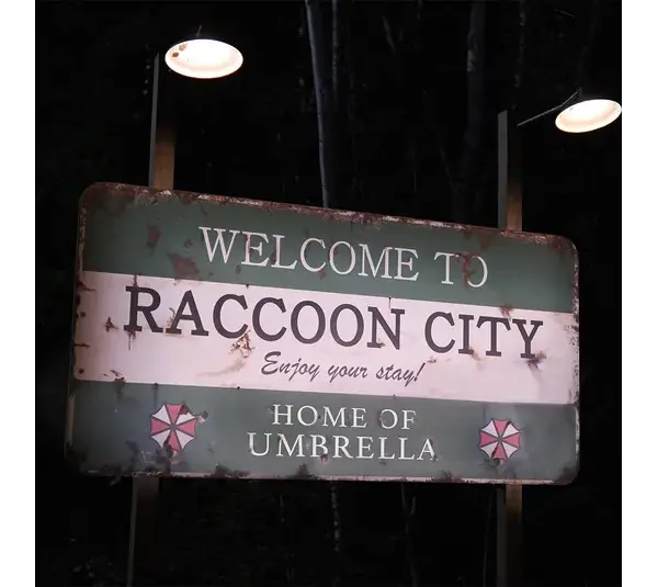 A weathered green sign lit by small lamps reads "Welcome to Raccoon City, Enjoy your stay!" and "Home of Umbrella." A foreboding nighttime forest background sets an eerie tone in Resident Evil.