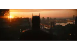 Silhouetted figure with pointed ears stands on a rooftop, gazing over a cityscape at sunset. Buildings and a river glow with warm light. Atmospheric mood. "The Batman: Part 2" is set for release in 2027.