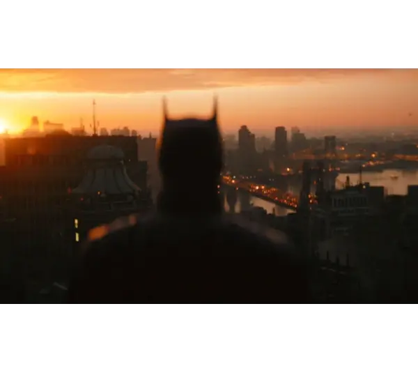 Silhouetted figure with pointed ears stands on a rooftop, gazing over a cityscape at sunset. Buildings and a river glow with warm light. Atmospheric mood. "The Batman: Part 2" is set for release in 2027.