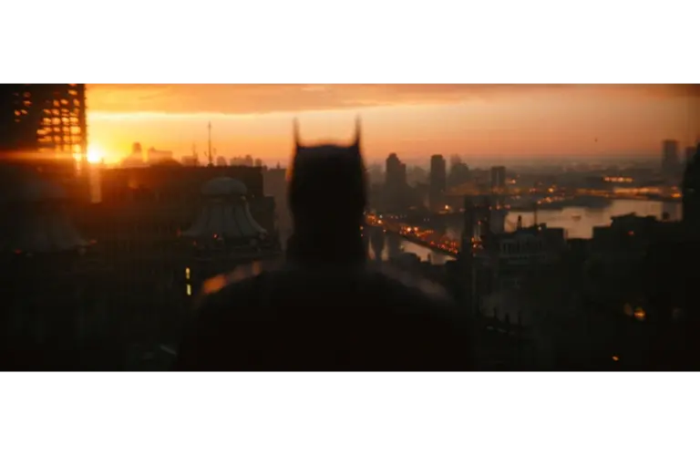 Silhouetted figure with pointed ears stands on a rooftop, gazing over a cityscape at sunset. Buildings and a river glow with warm light. Atmospheric mood. "The Batman: Part 2" is set for release in 2027.