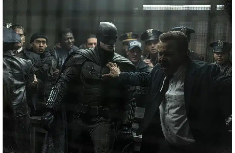 Batman stands amid police officers in a dimly lit room, holding back a yelling man in a suit. The scene is tense and chaotic.