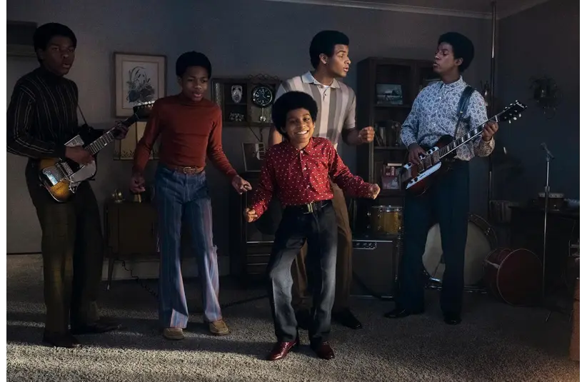 The cast of "Michael (2026)" perform enthusiastically in a living room, two playing guitars. The scene is lively and joyful, reflecting excitement and talent.