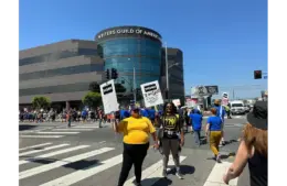 Protesters gather outside the WGA building, holding signs under a clear sky. The atmosphere is determined and unified.