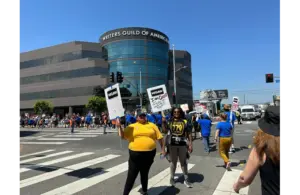 Protesters gather outside the WGA building, holding signs under a clear sky. The atmosphere is determined and unified.