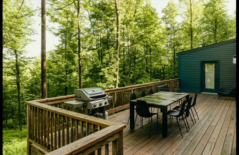 Wooden deck with a barbecue grill and a dining table set for eight. Surrounded by lush green trees, offering a serene, forested view.