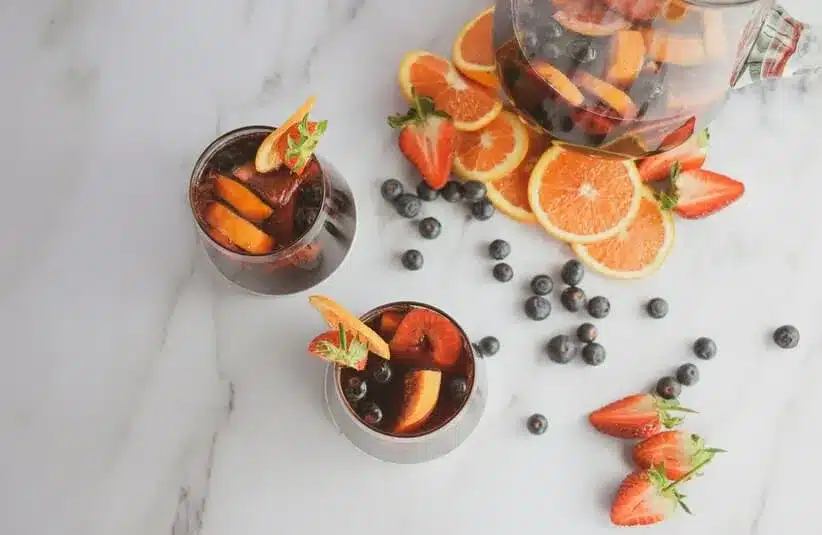 Image of two spring cocktails on a marble surface surrounded by sliced oranges, strawberries, and scattered blueberries, evoking a fresh, summery vibe.