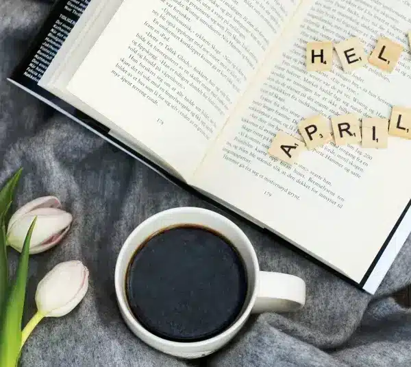 An open book with the words "Hello April" spread out across the pages, with a cup of coffee and some tulips on a table.
