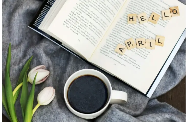 An open book with the words "Hello April" spread out across the pages, with a cup of coffee and some tulips on a table.