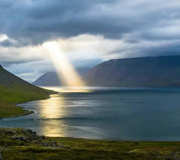 A beam of sunlight pierces through dark clouds, illuminating a serene fjord in Iceland and its surrounding green mountains, creating a peaceful and mystical atmosphere. The Nature of God. Heaven.