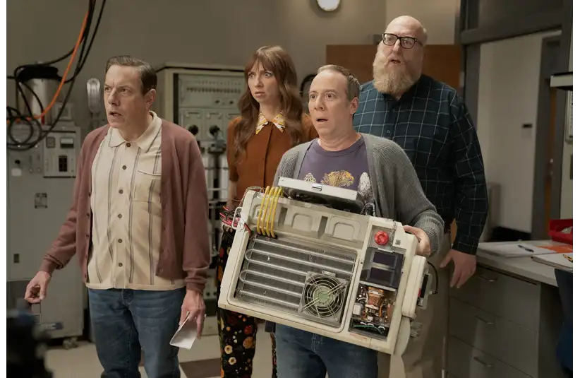 The characters of "Stuart Fails To Save The Universe" stand in a laboratory, looking surprised. One holds a homemade device with wires and circuits. The scene conveys curiosity and intrigue.