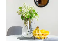 A vase with white flowers and green leaves sits on a round table next to a wire fruit basket filled with bananas and oranges, creating a fresh, inviting atmosphere. Spring decorating