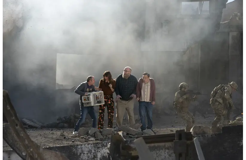The characters of "Stuart Fails To Save The Universe" huddle together in a smoky, debris-filled urban setting. Two armed soldiers guard them. The scene conveys tension and urgency.
