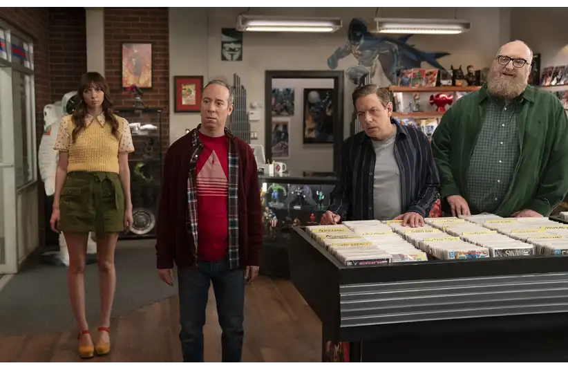 The characters of "Stuart Fails To Save The Universe" stand in a comic book store, looking perplexed. Shelves of comics and superhero posters adorn the background.