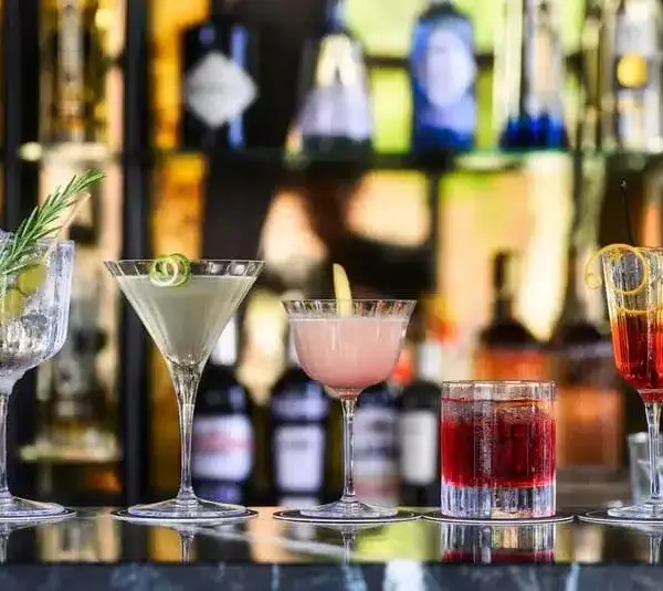 Image of a row of five spring cocktails on a bar counter, each in a distinct glass. Garnishes include rosemary, olives, citrus peels, and vibrant, colorful tones.