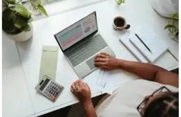 A woman works at a desk with a laptop displaying graphs, papers, a calculator, a notebook with a pencil, a coffee cup, and green plants. The scene conveys a budgeting exercise.