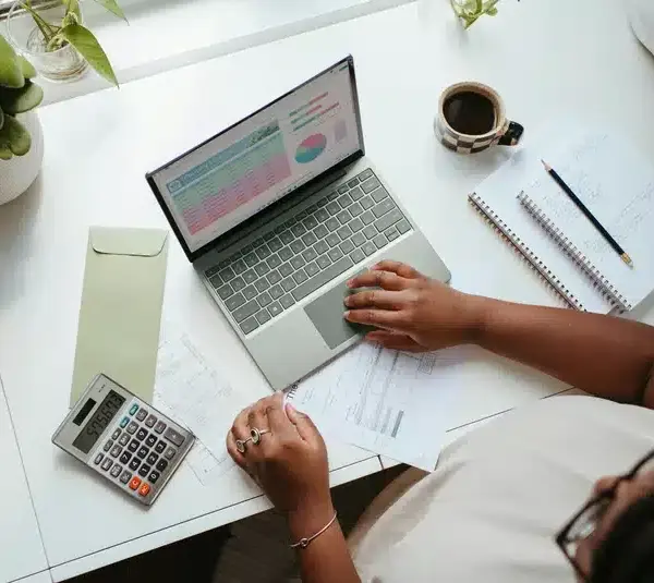 A woman works at a desk with a laptop displaying graphs, papers, a calculator, a notebook with a pencil, a coffee cup, and green plants. The scene conveys a budgeting exercise.