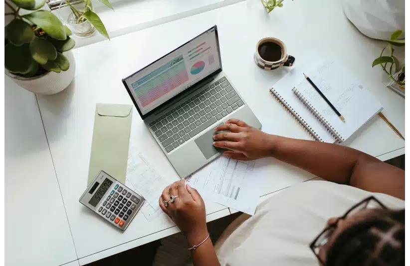 A woman works at a desk with a laptop displaying graphs, papers, a calculator, a notebook with a pencil, a coffee cup, and green plants. The scene conveys a budgeting exercise.