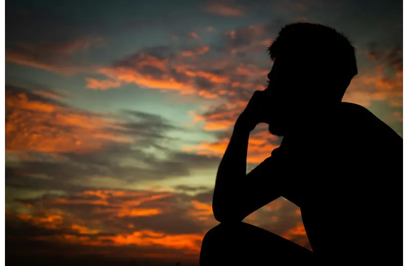Silhouette of a person deep in thought against a vibrant sunset sky filled with orange and blue hues, evoking a contemplative and serene mood.