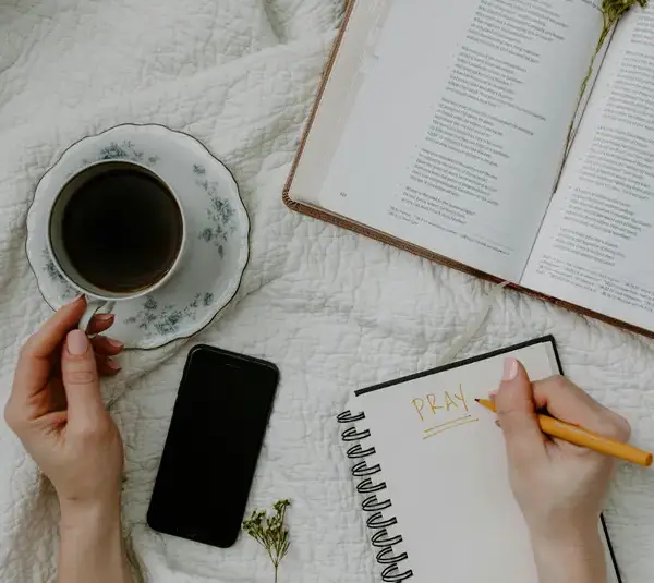 A person writes "Pray" in a notebook on a quilted surface for a daily devotional, next to an open book, a cup of coffee on a saucer, and a smartphone. The scene is calm and reflective.