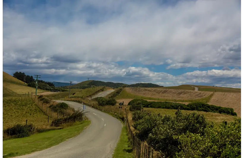 A winding rural road curves through a lush, green landscape with rolling hills under a cloudy sky, suggesting a peaceful, scenic journey.