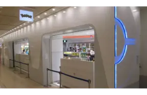 A sleek, modern Pokémon Centers exterior with blue neon accents and a white facade resembling a train. Interior shelves display colorful merchandise. Bright, clean ambiance. The Pokémon Company