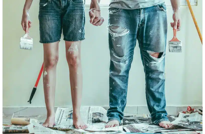 Two people stand barefoot on newspaper-covered floor, with paint on their shorts and jeans. They hold paintbrushes, conveying teamwork and creativity.