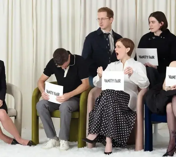 A group of six seated people holding "Vanity Fair" cards. One person expresses surprise, others are smiling or looking serious. White curtain backdrop.