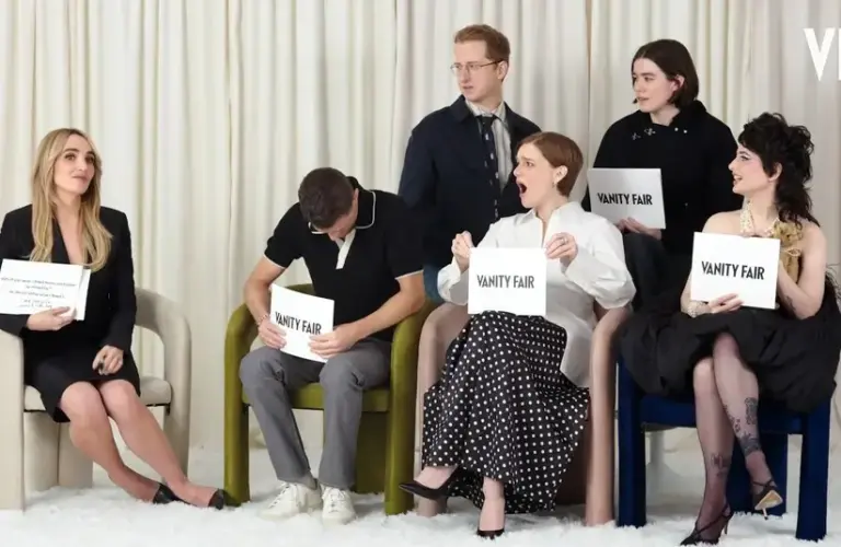 A group of six seated people holding "Vanity Fair" cards. One person expresses surprise, others are smiling or looking serious. White curtain backdrop.