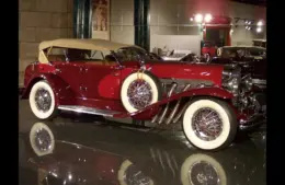 A shiny vintage Duesenberg convertible car in deep red with white-wall tires and chrome detailing is displayed in a museum setting, evoking elegance and nostalgia.