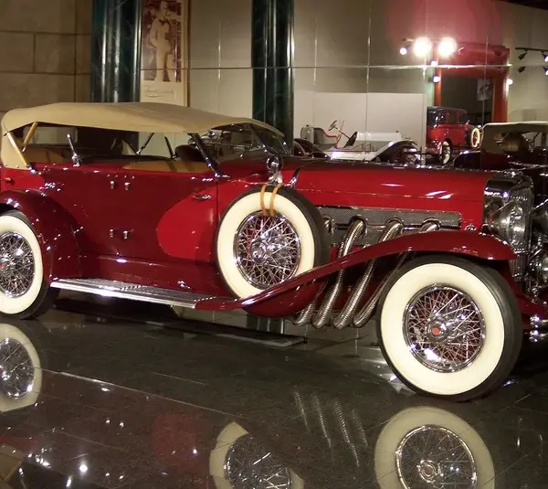 A shiny vintage Duesenberg convertible car in deep red with white-wall tires and chrome detailing is displayed in a museum setting, evoking elegance and nostalgia.