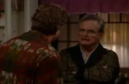 screenshot of Bonnie Bartlett husband William Dainels in the TV series "Boy Meets World" with glasses, arms crossed, faces another person wearing a patterned shirt in a dimly lit room. The tone is serious and contemplative.