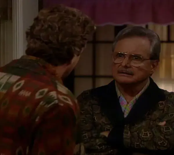 screenshot of Bonnie Bartlett husband William Dainels in the TV series "Boy Meets World" with glasses, arms crossed, faces another person wearing a patterned shirt in a dimly lit room. The tone is serious and contemplative.