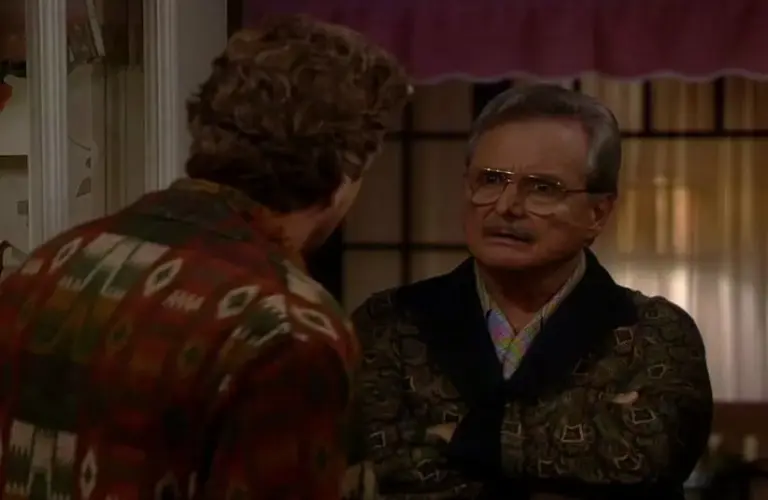 screenshot of Bonnie Bartlett husband William Dainels in the TV series "Boy Meets World" with glasses, arms crossed, faces another person wearing a patterned shirt in a dimly lit room. The tone is serious and contemplative.