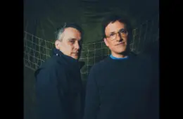 Anthony Russo and Joe Russo, directors of Avengers: Doomsday, stand in front of a green, net-like backdrop. One wears a black jacket, the other a dark sweater, both with neutral expressions.