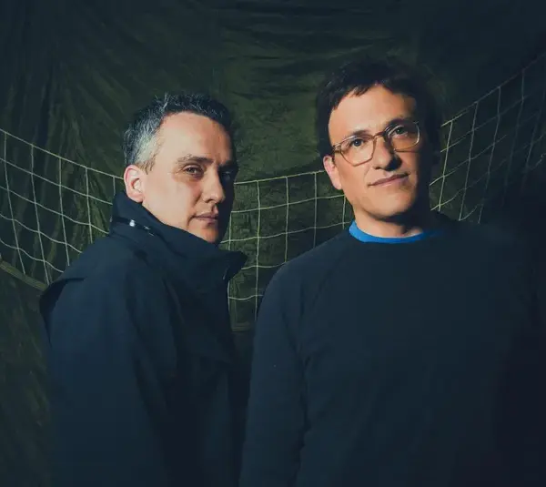 Anthony Russo and Joe Russo, directors of Avengers: Doomsday, stand in front of a green, net-like backdrop. One wears a black jacket, the other a dark sweater, both with neutral expressions.