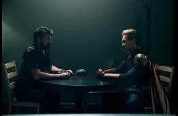 Karl Urban (Billy Butcher) and Antony Starr (Homelander) in The Boys sit across from each other at a small table in a dimly lit room, one in casual attire, the other in superhero costume, creating a tense atmosphere.
