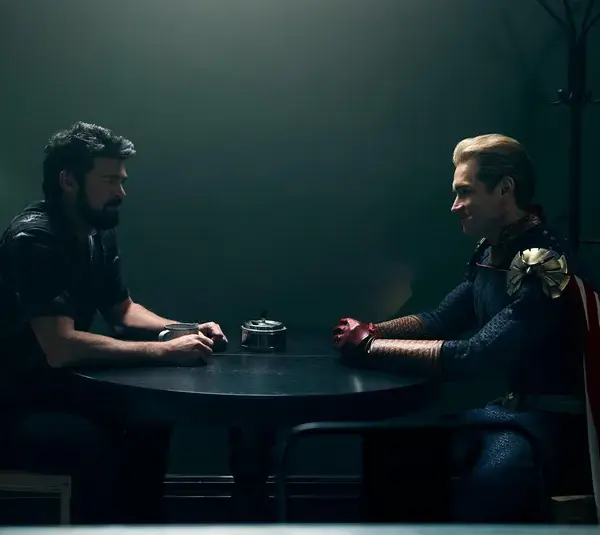 Karl Urban (Billy Butcher) and Antony Starr (Homelander) in The Boys sit across from each other at a small table in a dimly lit room, one in casual attire, the other in superhero costume, creating a tense atmosphere.