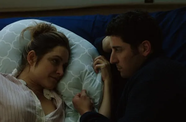 Jason Biggs and Jenny Mollen lie close, gazing at each other tenderly, holding hands. They're on a patterned pillow, exuding a calm and intimate atmosphere.