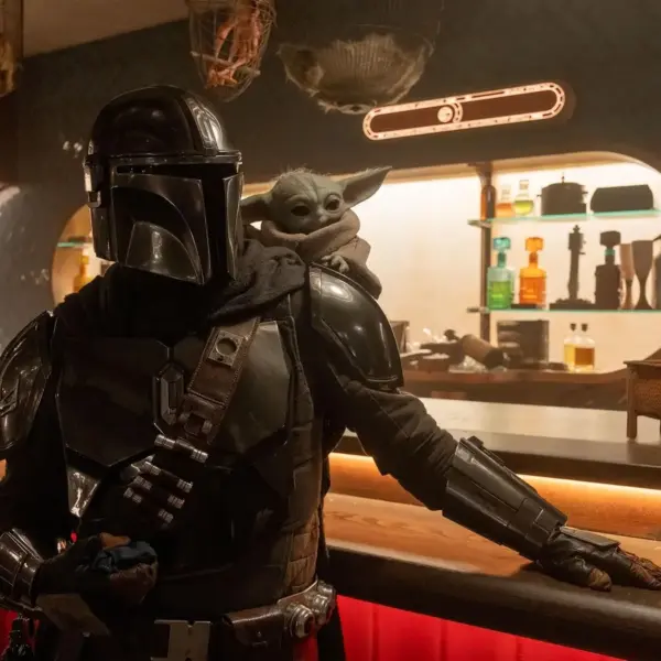 Pedro Pascal in "Star Wars: The Mandalorian and Grogu (2026)" stands confidently at a dimly lit bar, carrying a small, green, big-eared creature on their shoulder. The setting is futuristic and warm.