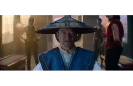 Tadanobu Asano in Mortal Kombat 2 (2026), in a blue robe and wide-brimmed hat, stares intently, his eyes glowing blue. Three people stand behind him in a dimly lit room, creating a tense atmosphere.