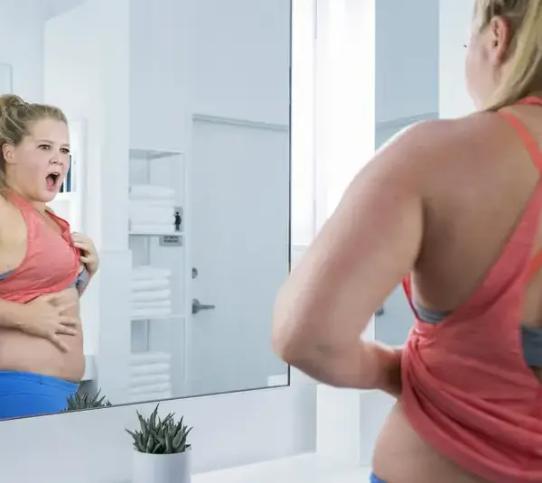 Amy Schumer, in "I Feel Pretty (2018)," in a pink tank top, stands in a bright bathroom, looking surprised at her reflection while pulling her shirt up to expose her stomach. Her expression conveys curiosity and shock.