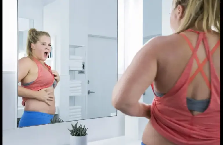 Amy Schumer, in "I Feel Pretty (2018)," in a pink tank top, stands in a bright bathroom, looking surprised at her reflection while pulling her shirt up to expose her stomach. Her expression conveys curiosity and shock.