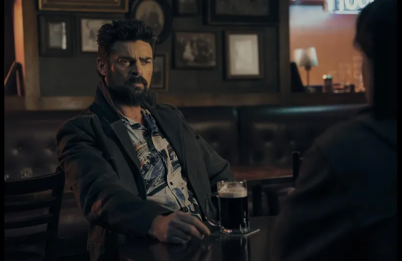 Karl Urban in The Boys (2019) sits in a dimly lit pub, holding a mug of dark beer. Framed pictures adorn the wall behind him, adding a nostalgic tone.