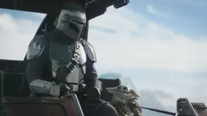 A helmeted figure in shiny armor sits beside a small, green creature with large ears. They're in a futuristic cockpit, with a serene sky in the background.
