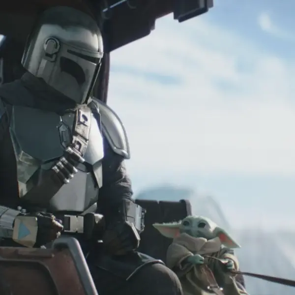 A helmeted figure in shiny armor sits beside a small, green creature with large ears. They're in a futuristic cockpit, with a serene sky in the background.