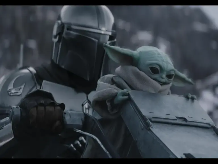 Pedro Pascal in Star Wars: The Mandalorian and Grogu (2026) rides a speeder bike with a small green creature bundled in a robe. The background shows a blurred, wintry landscape.