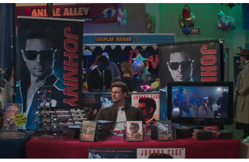 Karl Urban in Mortal Kombat 2 (2026) sits at a promotional booth with "Johnny" themed posters. He's surrounded by DVDs, sunglasses, and a TV display. The ambiance is lively and colorful.